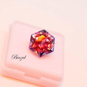 Puede incluir: Una piedra preciosa hexagonal facetada que muestra un degradado vibrante de tonos rojos, naranjas y morados. La piedra descansa sobre una base cuadrada de color rosa claro. La palabra Binzal est&aacute; impresa en negro en el lado izquierdo.