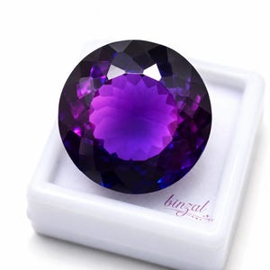 May include: A large, round amethyst gemstone with a deep purple hue, set in a white plastic case. The gem's facets reflect light, creating a brilliant display of color. The word "binzal" is printed in the lower right corner.