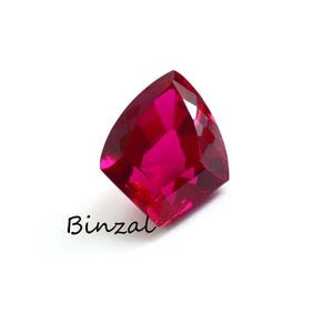 May include: A vibrant, deep red, cushion-cut gemstone with multiple facets that reflect light. The stone has a distinct, saturated color and a polished surface. The word Binzal is printed in black script text below the gemstone on a white background.
