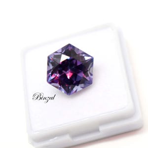 Puede incluir: Una piedra preciosa hexagonal facetada con tonos p&uacute;rpuras y violetas profundos, que muestra cortes geom&eacute;tricos n&iacute;tidos y reflejos de luz. La piedra est&aacute; colocada sobre un estuche cuadrado blanco con el texto Binzal en cursiva negra.