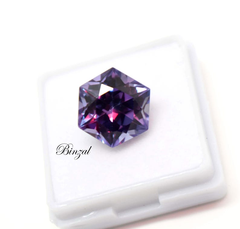 Puede incluir: Una piedra preciosa hexagonal facetada con tonos p&uacute;rpuras y violetas profundos, que muestra cortes geom&eacute;tricos n&iacute;tidos y reflejos de luz. La piedra est&aacute; colocada sobre un estuche cuadrado blanco con el texto Binzal en cursiva negra.