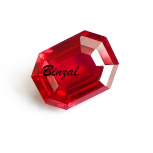 以下が含まれることがあります： 「Binzal」の筆記体文字が入った、ファセットカットの長方形の濃い赤色の宝石。宝石には光を反射する複数のファセットがあり、鮮やかな外観を作り出しています。宝石は白い背景に配置されています。