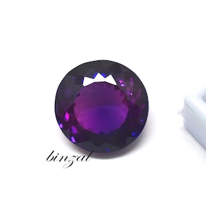 May include: A round, faceted gemstone with a deep purple hue, transitioning to lighter shades in the center. The gem is set against a white background, with the word "binzal" in cursive script.