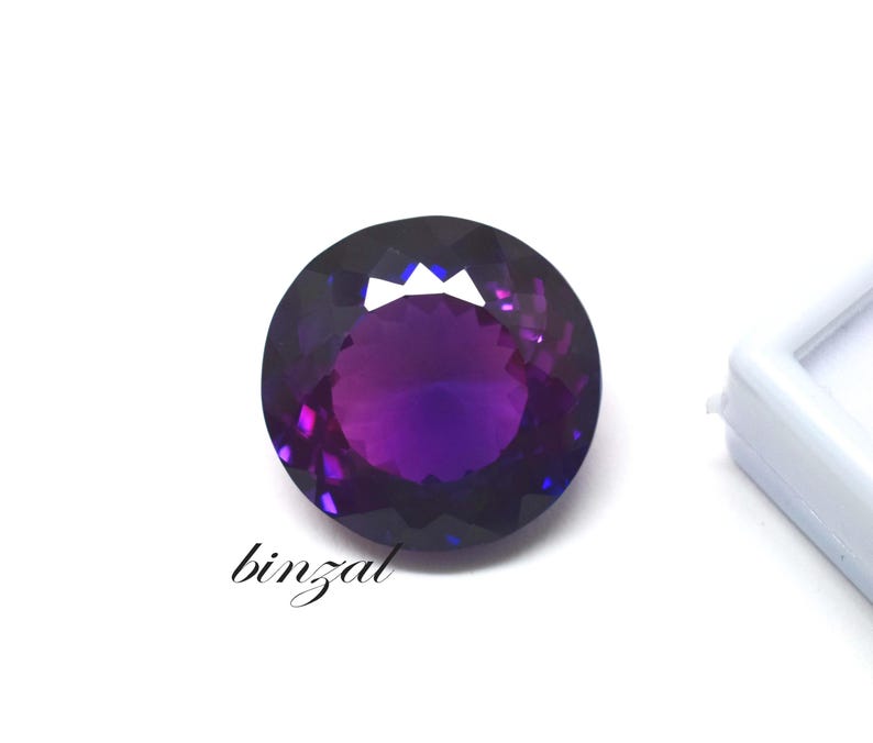 May include: A round, faceted gemstone with a deep purple hue, transitioning to lighter shades in the center. The gem is set against a white background, with the word "binzal" in cursive script.