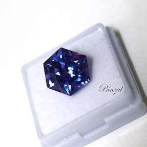 Puede incluir: Una piedra preciosa hexagonal facetada con tonos azul profundo y violeta, que muestra un corte brillante con m&uacute;ltiples superficies reflectantes. La piedra est&aacute; colocada sobre una base de exhibici&oacute;n cuadrada blanca con la palabra Binzal en negro.