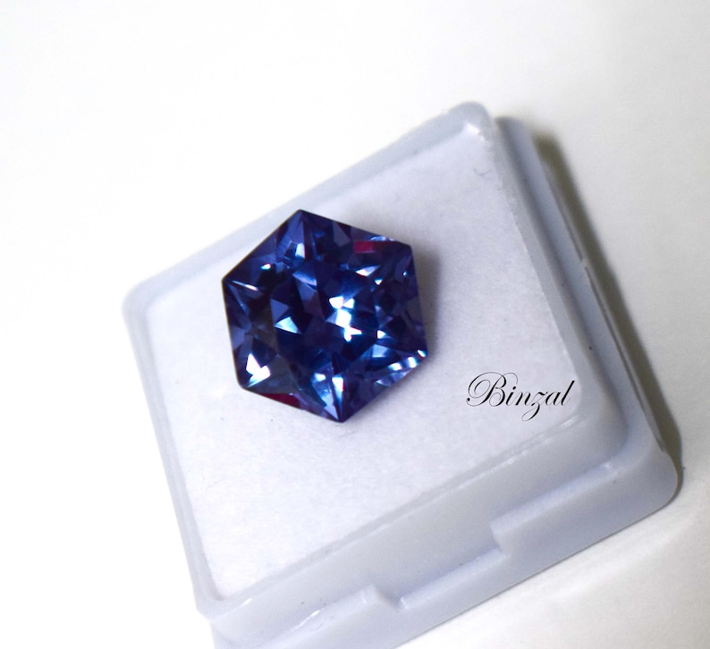 Puede incluir: Una piedra preciosa hexagonal facetada con tonos azul profundo y violeta, que muestra un corte brillante con m&uacute;ltiples superficies reflectantes. La piedra est&aacute; colocada sobre una base de exhibici&oacute;n cuadrada blanca con la palabra Binzal en negro.