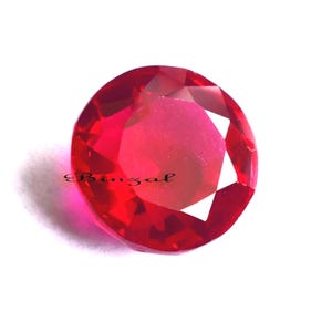 Op de afbeelding: Een gefacetteerde, ronde, helderrode edelsteen. De edelsteen heeft een hoge mate van helderheid en schittering en reflecteert licht. Het woord "Binzal" staat op de edelsteen. De edelsteen staat tegen een witte achtergrond.