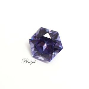 Puede incluir: Una piedra preciosa morada de forma pentagonal con facetas y un corte brillante que refleja la luz. La piedra muestra tonos violeta intenso y lavanda m&aacute;s claro en toda su superficie. La palabra Binzal est&aacute; impresa en texto negro.
