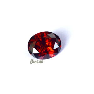 May include: An oval-shaped, faceted gemstone with a deep red color. The gem has a polished surface that reflects light, creating a sparkling effect. The word "Binzal" is printed on the bottom left of the image.