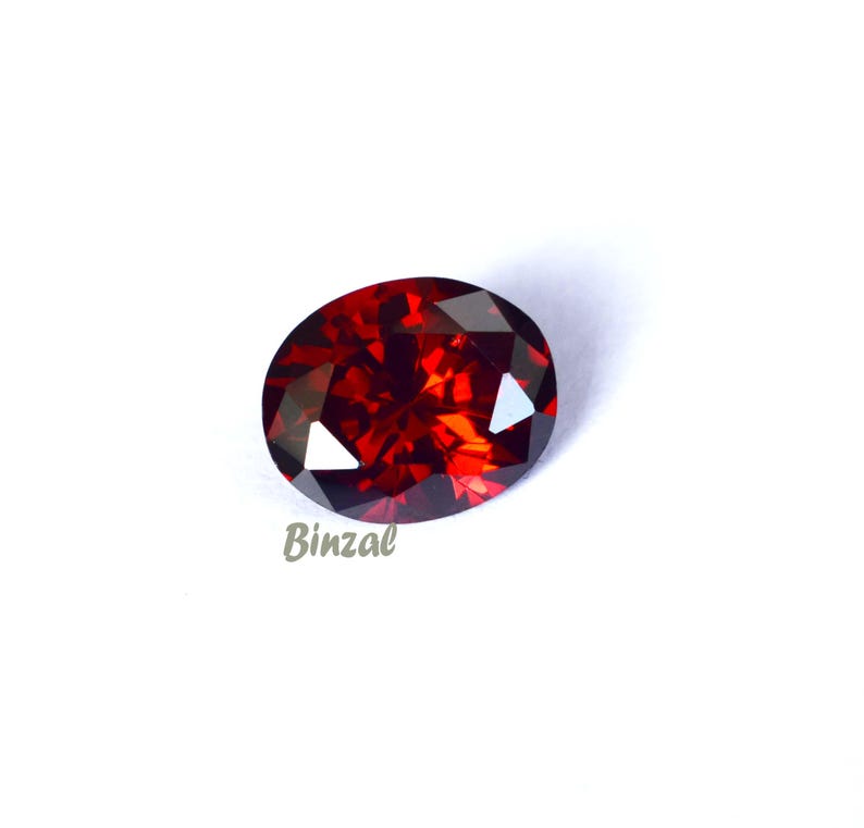 May include: An oval-shaped, faceted gemstone with a deep red color. The gem has a polished surface that reflects light, creating a sparkling effect. The word "Binzal" is printed on the bottom left of the image.
