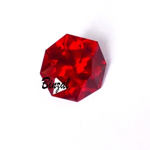 Puede incluir: Una gema facetada, octogonal, de color rojo intenso. La gema tiene múltiples superficies reflectantes, que captan y dispersan la luz. La palabra "Binzal" está impresa en la gema. La gema está sobre un fondo blanco.