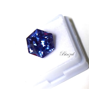 Puede incluir: Una piedra preciosa azul hexagonal facetada con tonos violetas profundos, que muestra una superficie reflectante brillante. La piedra est&aacute; colocada sobre una base blanca con el texto Binzal impreso en una elegante fuente cursiva negra.