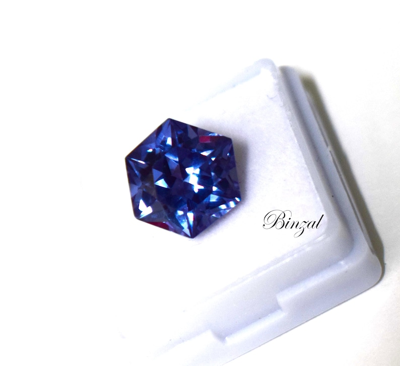 Puede incluir: Una piedra preciosa azul hexagonal facetada con tonos violetas profundos, que muestra una superficie reflectante brillante. La piedra est&aacute; colocada sobre una base blanca con el texto Binzal impreso en una elegante fuente cursiva negra.