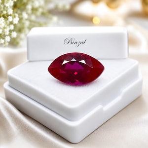 May include: A marquise-cut, deep red gemstone rests in a white jewelry box. The gem is faceted, catching the light and displaying a vibrant color. The box is open, revealing a soft, white interior. The word "Binzal" is printed on the box.
