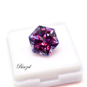 Puede incluir: Una piedra preciosa hexagonal facetada que muestra tonos vibrantes de p&uacute;rpura, magenta y azul profundo. La piedra est&aacute; colocada sobre una caja de exhibici&oacute;n cuadrada blanca con el texto Binzal impreso en fuente cursiva negra.