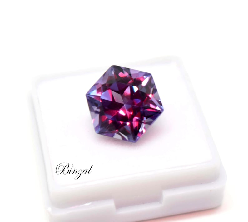 Puede incluir: Una piedra preciosa hexagonal facetada que muestra tonos vibrantes de p&uacute;rpura, magenta y azul profundo. La piedra est&aacute; colocada sobre una caja de exhibici&oacute;n cuadrada blanca con el texto Binzal impreso en fuente cursiva negra.