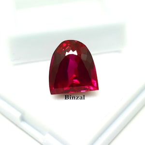 Può includere: Una gemma sfaccettata, rosso intenso, dalla forma unica, su sfondo bianco. La gemma ha una superficie lucida e un colore vibrante. La parola "Binzal" è stampata sul fondo della gemma.