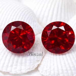 Könnte beinhalten: Zwei runde, facettierte, tiefrote Edelsteine. Die Edelsteine sind vor einem Hintergrund aus weißem, strukturiertem Material platziert. Das Wort "Binzal" ist in kursiver Schrift unter den Edelsteinen gedruckt.