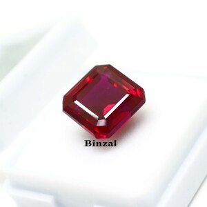 May include: A faceted, square-cut ruby gemstone with a deep red hue. The gem is set against a white background. The word "Binzal" is printed in black text below the gemstone.