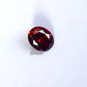 May include: An oval-shaped, faceted, deep red gemstone. The gem has multiple reflective surfaces, creating bright highlights. The word "Binzae" is printed in light green on the lower left side of the image. The gemstone is set against a white background.