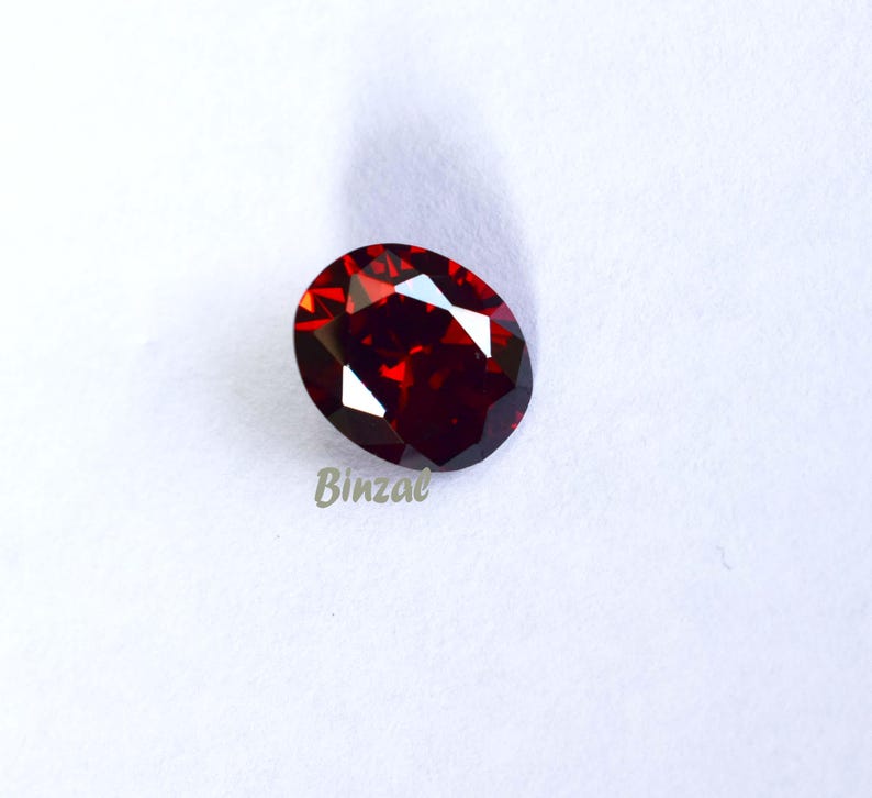 May include: An oval-shaped, faceted, deep red gemstone. The gem has multiple reflective surfaces, creating bright highlights. The word "Binzae" is printed in light green on the lower left side of the image. The gemstone is set against a white background.