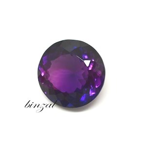 May include: A faceted, round amethyst gemstone with a deep purple hue and lighter purple and blue highlights. The word "binzal" is written in cursive on the lower left side of the image. The gemstone is set against a white background.