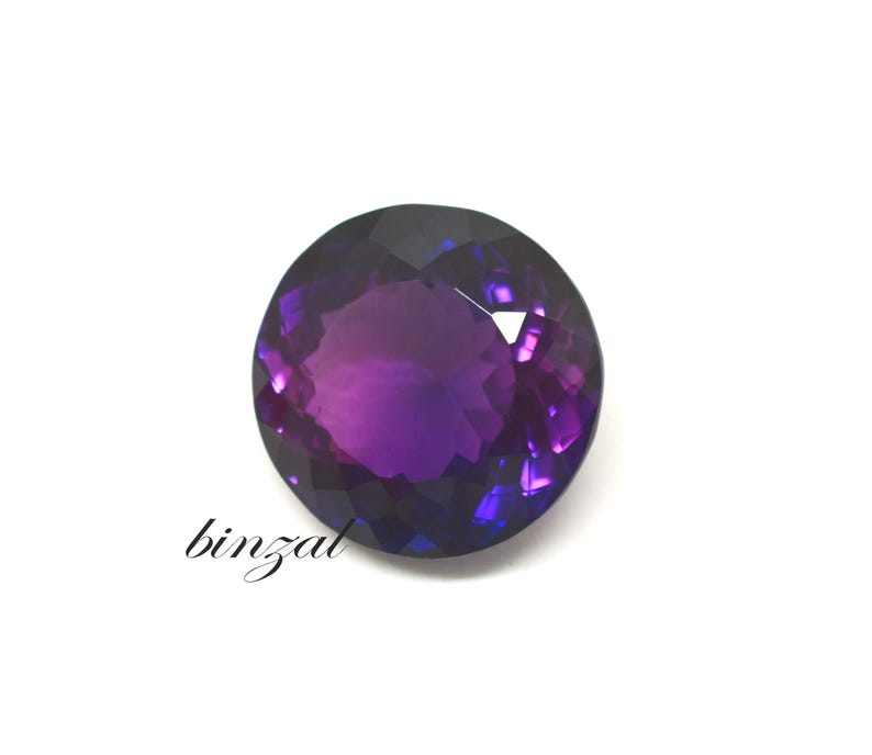 May include: A faceted, round amethyst gemstone with a deep purple hue and lighter purple and blue highlights. The word "binzal" is written in cursive on the lower left side of the image. The gemstone is set against a white background.