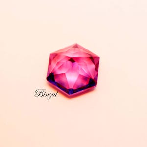 Puede incluir: Una piedra preciosa hexagonal facetada que muestra un degradado vibrante de tonos magenta intenso y violeta. La piedra presenta cortes geom&eacute;tricos precisos que crean un patr&oacute;n de reflexi&oacute;n de luz en forma de estrella. La palabra Binzal est&aacute; impresa en negro.