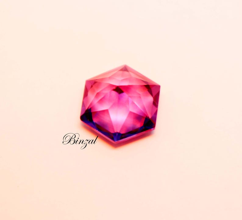 Puede incluir: Una piedra preciosa hexagonal facetada que muestra un degradado vibrante de tonos magenta intenso y violeta. La piedra presenta cortes geom&eacute;tricos precisos que crean un patr&oacute;n de reflexi&oacute;n de luz en forma de estrella. La palabra Binzal est&aacute; impresa en negro.