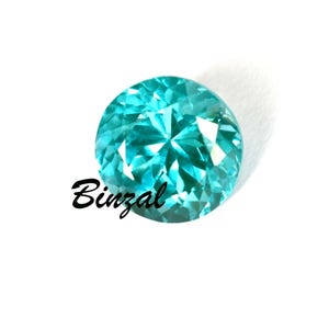 May include: A round, faceted, teal gemstone with a brilliant cut. The gem is set against a white background, with the word "Binzal" in black script on the lower left side. The gem's facets reflect light, creating a sparkling effect.