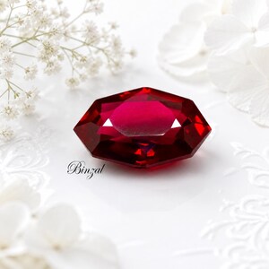 May include: A faceted, deep red gemstone with a geometric shape. The gem is set against a white background with delicate white flowers and the word "Binzal" in cursive script.