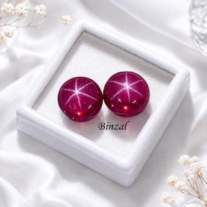 Pode incluir: Dois rubis estrela redondos de cor magenta profunda com um asterismo distinto de seis raios. As pedras preciosas são exibidas dentro de uma caixa quadrada branca com forro interno branco. O texto Binzal está impresso em preto.