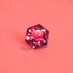 Puede incluir: Una piedra preciosa hexagonal facetada en un tono rojo magenta intenso con bordes geom&eacute;tricos afilados y una superficie reflectante brillante. La palabra Binzal est&aacute; impresa en una fuente cursiva negra debajo de la piedra sobre un fondo de color coral claro.