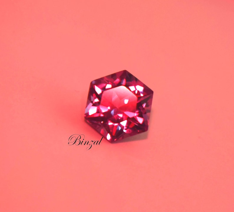 Puede incluir: Una piedra preciosa hexagonal facetada en un tono rojo magenta intenso con bordes geom&eacute;tricos afilados y una superficie reflectante brillante. La palabra Binzal est&aacute; impresa en una fuente cursiva negra debajo de la piedra sobre un fondo de color coral claro.