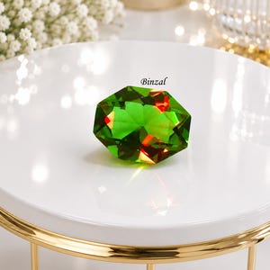 Può includere: Una gemma di cristallo verde smeraldo sfaccettata con una forma geometrica a più lati. Il cristallo presenta riflessi di luce interni e la parola Binzal è stampata in piccoli caratteri neri sopra il bordo superiore della pietra verde.
