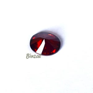 May include: A faceted, oval-shaped, deep red gemstone. The gem has a cut-out section on one side, revealing a white interior. The word "Binzal" is printed in a light brown color on the lower left side of the image.