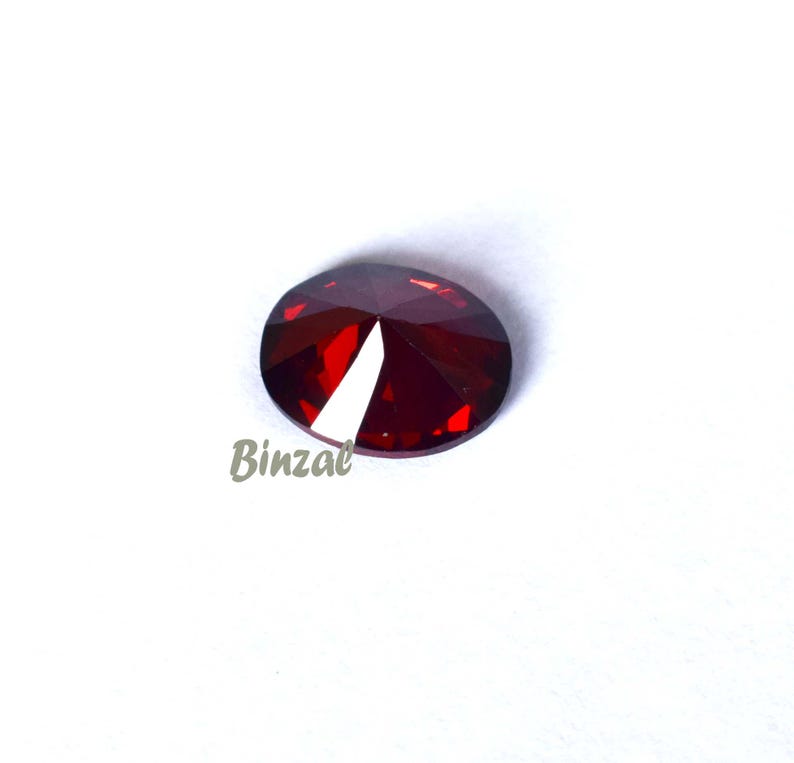 May include: A faceted, oval-shaped, deep red gemstone. The gem has a cut-out section on one side, revealing a white interior. The word "Binzal" is printed in a light brown color on the lower left side of the image.