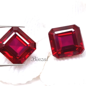 May include: Two radiant-cut, deep red gemstones. One is held by tweezers, the other rests beside it. The gems have a square shape with beveled edges, reflecting light. The background is white. The word "Binzal" is visible.