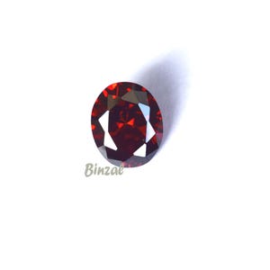 May include: A faceted, oval-cut, deep red gemstone. The gem has a brilliant shine and is set against a white background. The word "Binzae" is printed in the lower left corner.