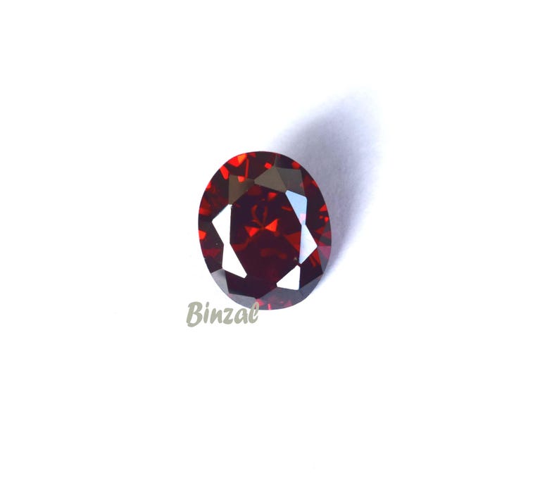 May include: A faceted, oval-cut, deep red gemstone. The gem has a brilliant shine and is set against a white background. The word "Binzae" is printed in the lower left corner.