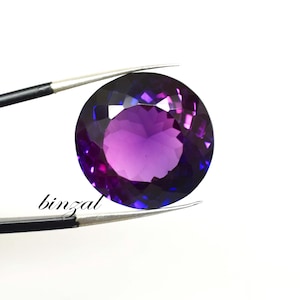 May include: A round, faceted gemstone with a gradient of purple and blue hues. The gem is held by a pair of tweezers against a white background. The word "binzal" is printed in black cursive font.