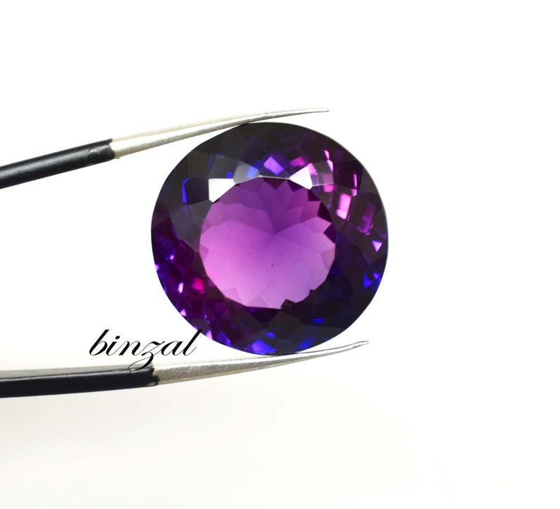 May include: A round, faceted gemstone with a gradient of purple and blue hues. The gem is held by a pair of tweezers against a white background. The word "binzal" is printed in black cursive font.