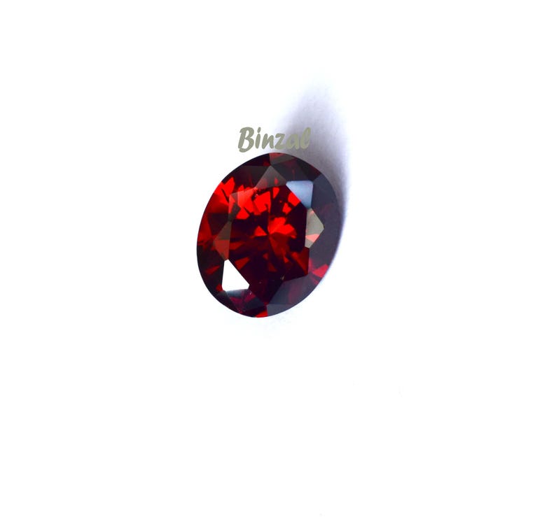 May include: A faceted, oval-shaped, deep red gemstone. The gem has a high degree of clarity and brilliance, reflecting light to create a sparkling effect. The word "Binzal" is printed in light green on the top left of the gem.