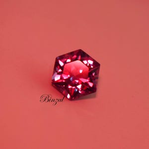 Puede incluir: Una piedra preciosa hexagonal facetada de color magenta intenso con un corte brillante que refleja la luz. La piedra presenta bordes geom&eacute;tricos afilados y una superficie pulida. La palabra Binzal est&aacute; impresa en letra cursiva negra debajo de la piedra.