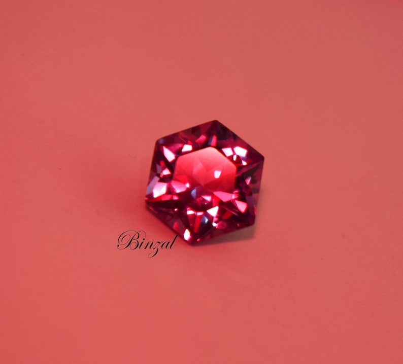 Puede incluir: Una piedra preciosa hexagonal facetada de color magenta intenso con un corte brillante que refleja la luz. La piedra presenta bordes geom&eacute;tricos afilados y una superficie pulida. La palabra Binzal est&aacute; impresa en letra cursiva negra debajo de la piedra.