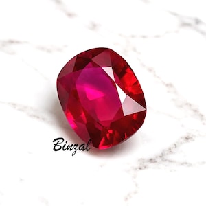 Peut inclure: Une pierre précieuse rubis facettée de forme ovale et de couleur rouge foncé. La gemme est posée sur une surface en marbre blanc. Le mot "Binzal" est écrit en cursive en bas de l'image.