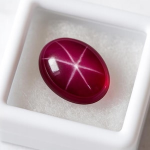 May include: A polished, oval-shaped ruby gemstone with a six-rayed star pattern. The gem is a deep red color and is set in a white display box. The star effect is created by light reflecting off of inclusions within the stone.