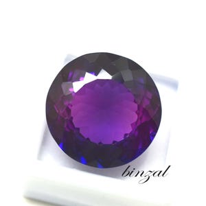 May include: A large, round, faceted gemstone with a deep purple hue, transitioning to lighter shades in the center. The gem is set against a white background, with the word "binzal" in cursive script.