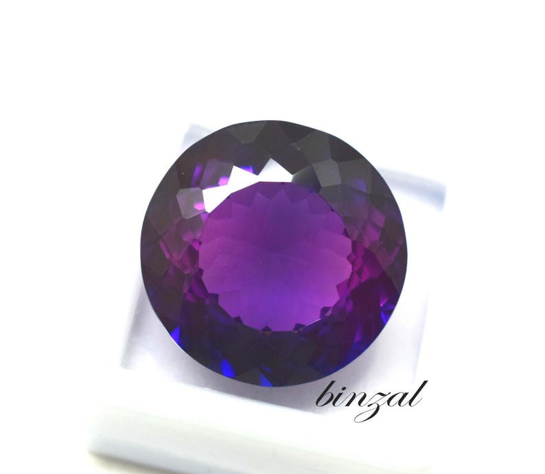 May include: A large, round, faceted gemstone with a deep purple hue, transitioning to lighter shades in the center. The gem is set against a white background, with the word "binzal" in cursive script.