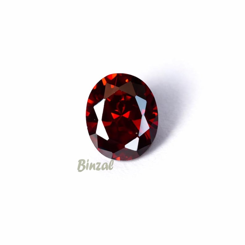 May include: A faceted, oval-shaped, deep red gemstone. The gem has a high degree of clarity and brilliance, reflecting light to create a sparkling effect. The word "Binzal" is printed in a cursive font on the lower left side of the image.